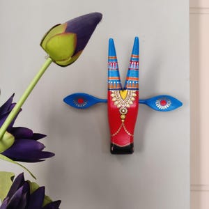 Pode incluir: Decora&ccedil;&atilde;o de parede de madeira pintada &agrave; m&atilde;o em forma de cabe&ccedil;a de touro. O touro tem um corpo vermelho, chifres e orelhas azuis e detalhes dourados e laranja. A pe&ccedil;a decorativa &eacute; montada em uma parede cinza claro, com flores roxas em primeiro plano.