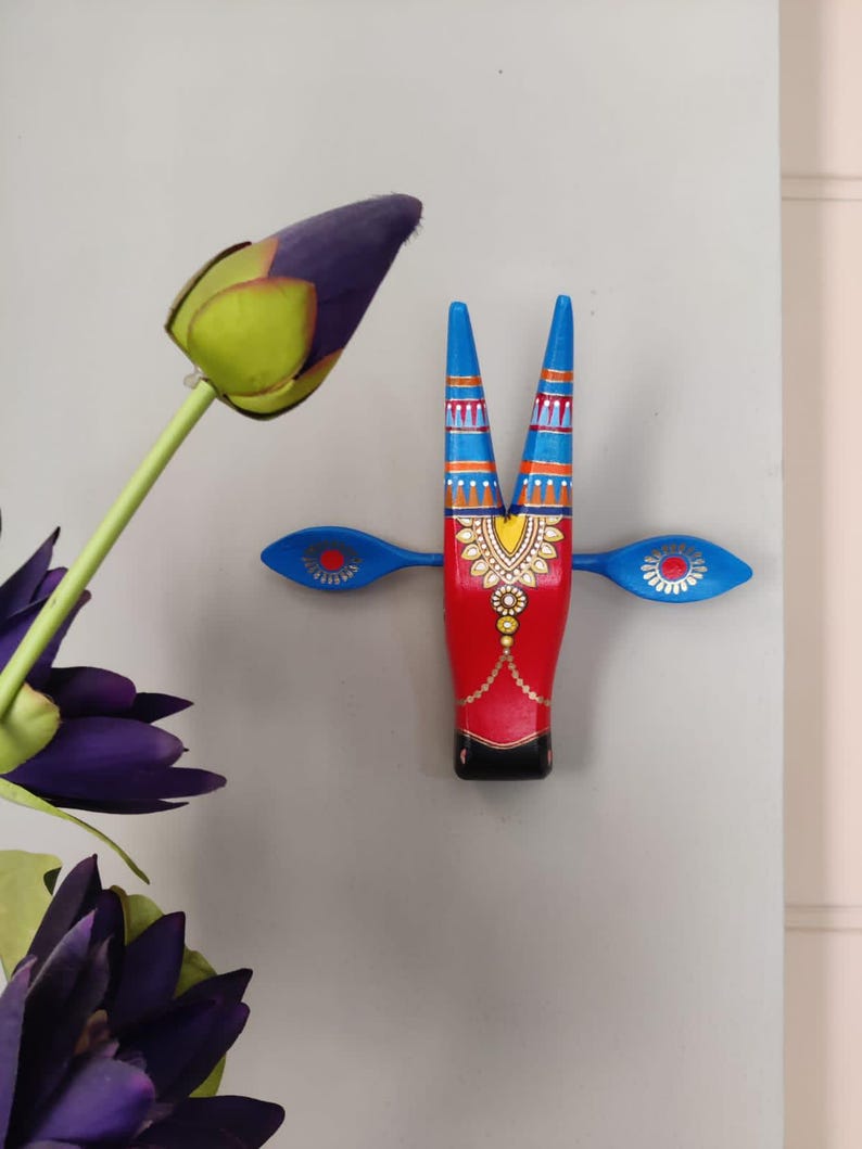 Pode incluir: Decora&ccedil;&atilde;o de parede de madeira pintada &agrave; m&atilde;o em forma de cabe&ccedil;a de touro. O touro tem um corpo vermelho, chifres e orelhas azuis e detalhes dourados e laranja. A pe&ccedil;a decorativa &eacute; montada em uma parede cinza claro, com flores roxas em primeiro plano.