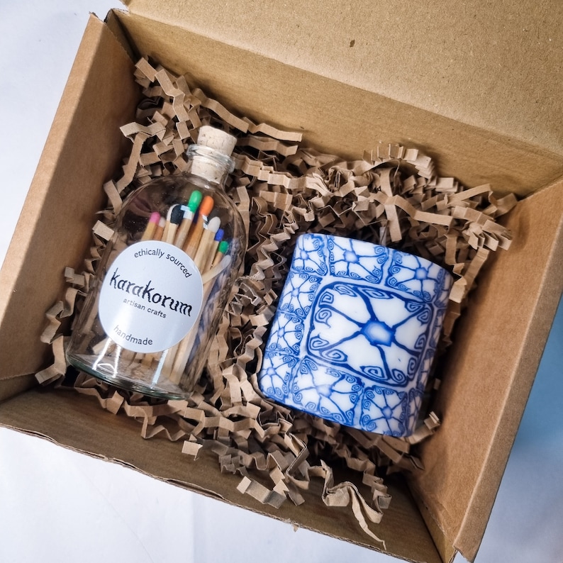 May include: A gift box filled with brown packing peanuts contains a glass jar of colorful matches and a blue and white ceramic candle. The jar has a white label that reads "ethically sourced Karakorum artisan crafts handmade".