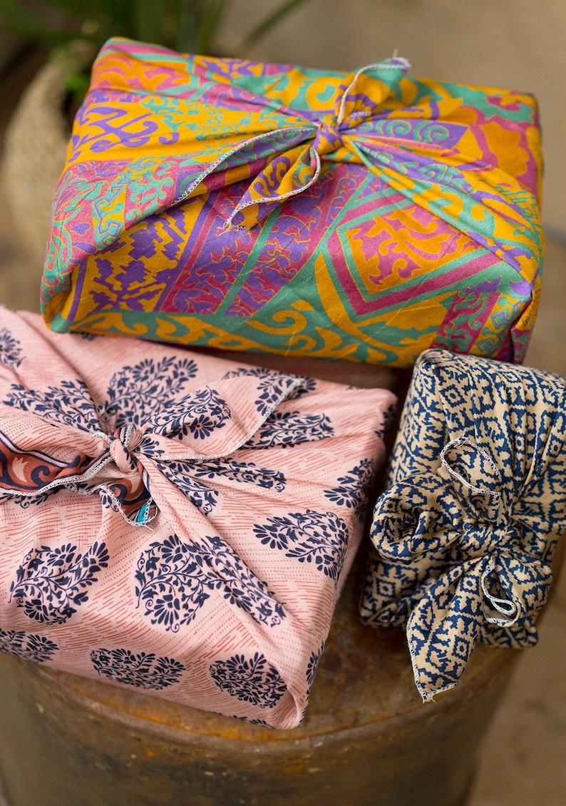 Pu&ograve; includere: Tre regali avvolti in tessuti colorati con diversi motivi. Il regalo in alto &egrave; avvolto in un tessuto giallo, rosa e viola con un motivo geometrico. Il regalo al centro &egrave; avvolto in un tessuto rosa con un motivo floreale blu. Il regalo in basso &egrave; avvolto in un tessuto blu e marrone con un motivo geometrico.