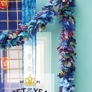 Op de afbeelding: Een blauwe stoffen slinger met stroken van bedrukte en effen textiel in diverse tinten blauw, paars en rood, gedrapeerd over een zwarte gordijnroede. De tekst 20 25 GIFT OF THE YEAR HIGHLY COMMENDED staat onderaan in het midden afgedrukt.