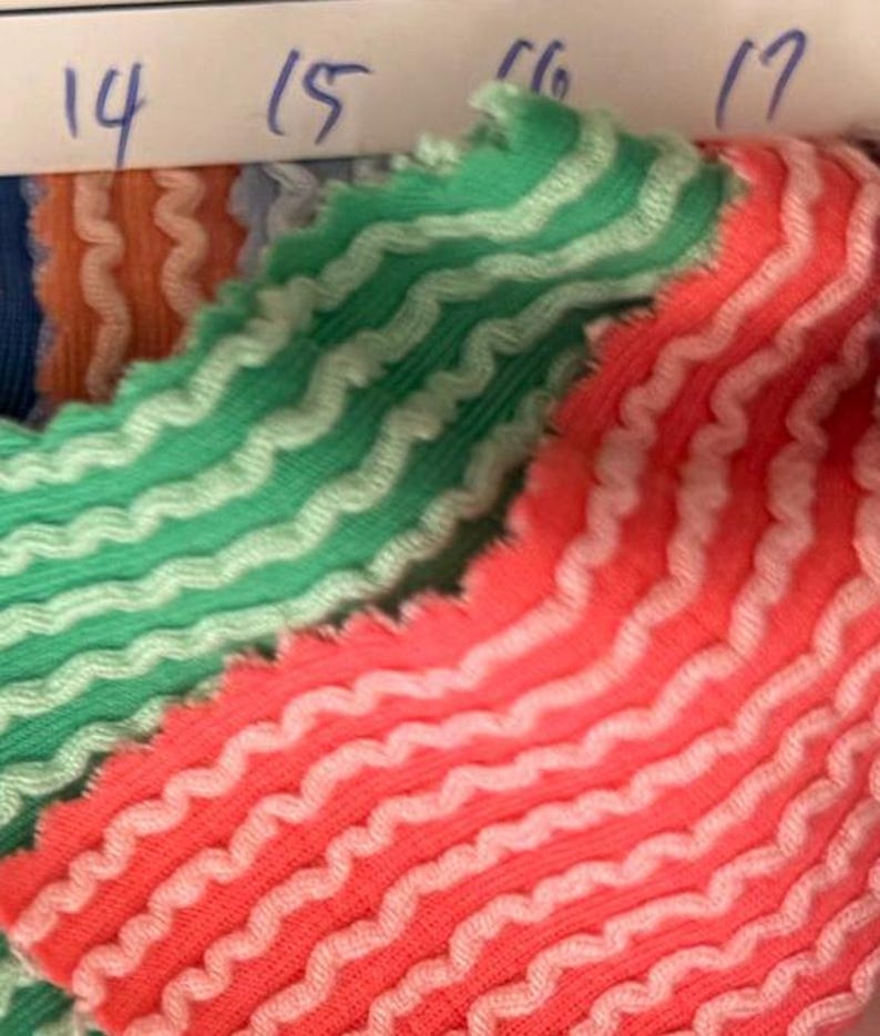 May include: Close-up of fabric samples with a wavy, textured design. The samples feature a repeating pattern of white lines on a green and coral background. The edges are scalloped. Numbers 14, 15, 16, and 17 are visible.