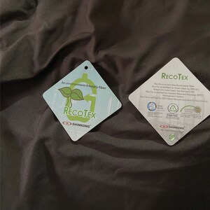 May include: Two white diamond-shaped tags with green and black text and logos. The top tag reads "An environment-friendly fibre" and "RECOTEX" with a green recycling symbol. The bottom tag reads "RECOTEX" and "SHINKONG" with a green recycling symbol and the text "RecoTex is an eco-friendly polyester fibre. RecoTex is certified as Green Mark by EPA of Taiwan and meet Oeko-Tex Standard 100. RecoTex is certified by Intertek Recycled Polyester (PET) Management System. RecoTex is certified by GRS Global Recycle Standard Control Union."