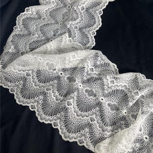 May include: A length of white lace fabric with a detailed floral pattern. The lace has a scalloped edge and is displayed against a dark background, highlighting its intricate design. This delicate fabric is suitable for various craft and sewing projects.