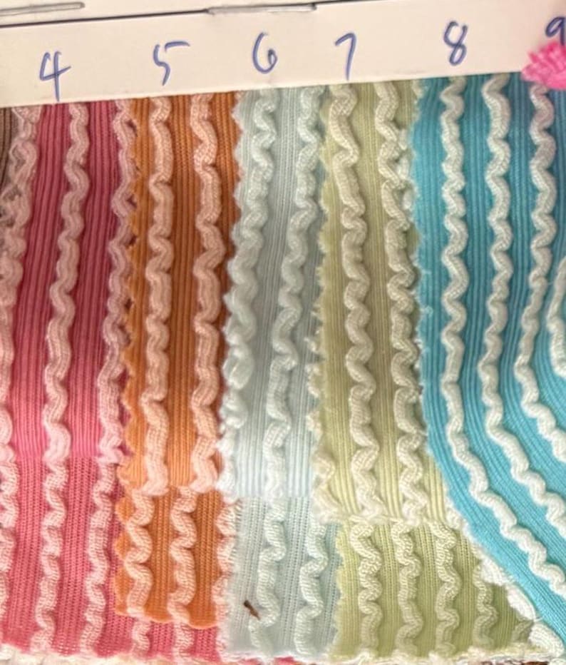 May include: Close-up of fabric swatches in various colors, including pink, orange, light blue, and yellow. Each swatch has a ribbed texture with a wavy, white trim along the edges. Numbers 4 through 9 are visible at the top.