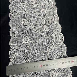 May include: Close-up of white floral lace fabric. The lace features a repeating pattern of large, stylized flowers with intricate details. A ruler is placed across the lace, showing measurements in centimeters.