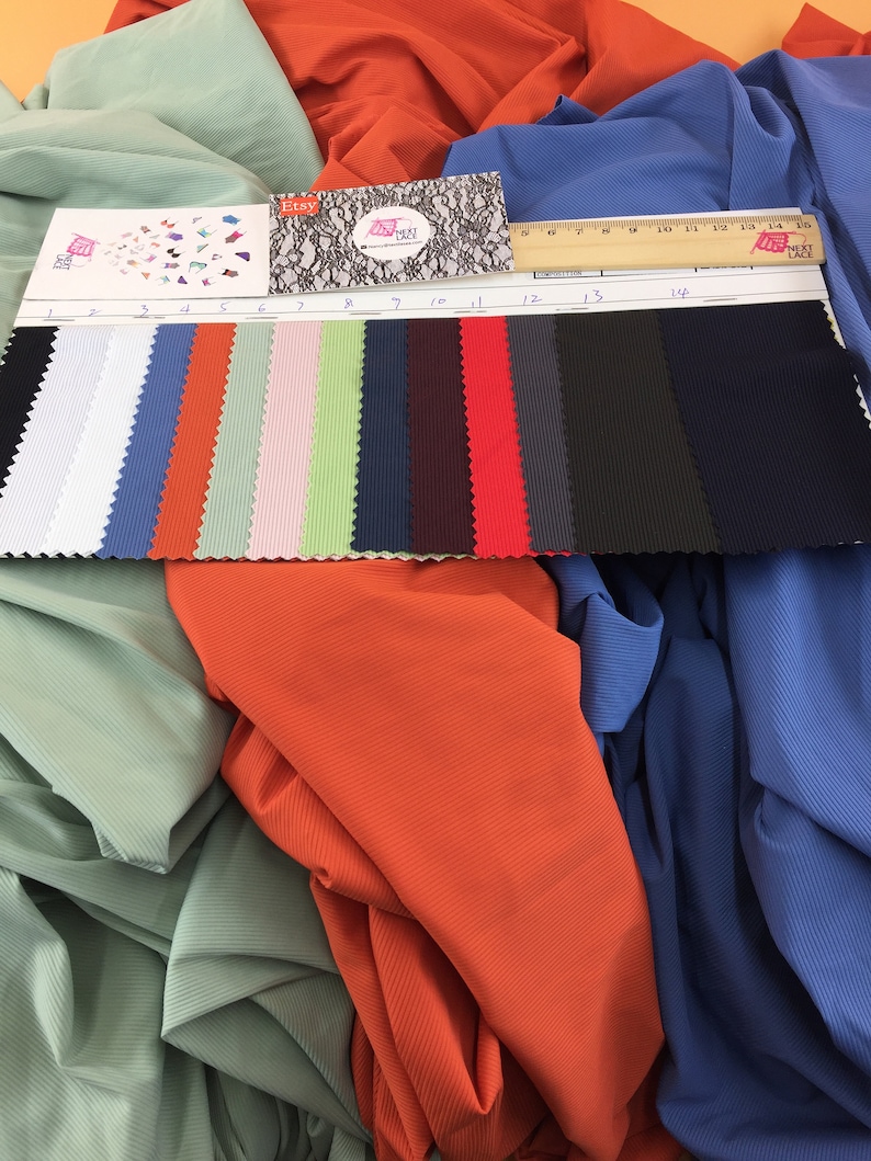May include: A fabric swatch card featuring fourteen distinct colors of ribbed material, numbered one through fourteen, displayed alongside a wooden ruler measuring 15 centimeters. The swatches include black, white, blue, orange, green, pink, and red.