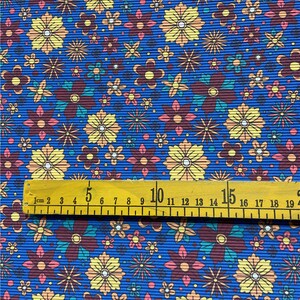 Op de afbeelding: Blauwe stof met een herhalend patroon van kleurrijke bloemen. Een houten liniaal met markeringen in centimeters ligt op de stof.
