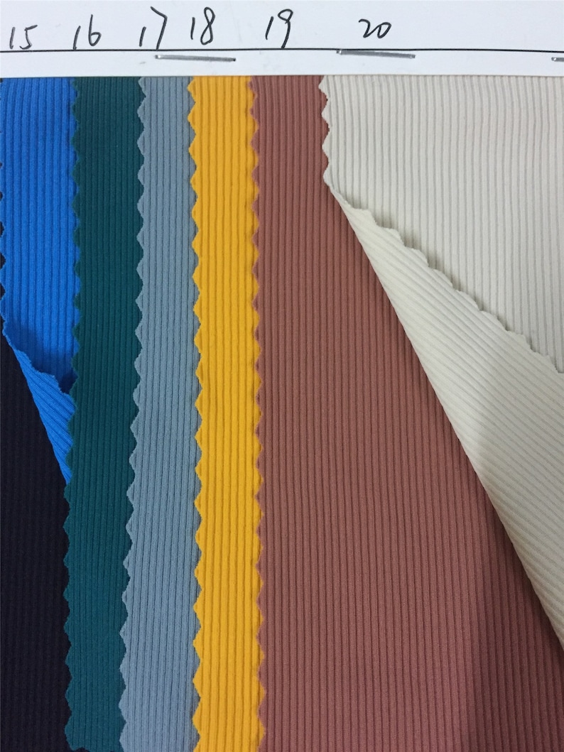 May include: A collection of ribbed fabric swatches in vertical strips of black, blue, dark teal, grey, yellow, terracotta, and cream. The top edge features a white paper header with the handwritten numbers 15, 16, 17, 18, 19, and 20.