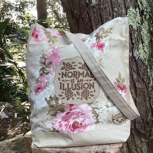 May include: Beige tote bag with a floral pattern in pink and white. The bag features a rectangular label with the text "NORMAL IS AN ILLUSION". The bag has a long strap and is made of canvas-like material.