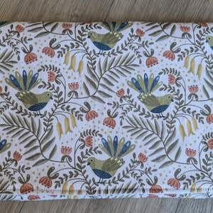 May include: A rectangular, folded fabric item featuring a repeating pattern of birds, flowers, and foliage in green, blue, and orange on a white background. Secured with two orange buttons.