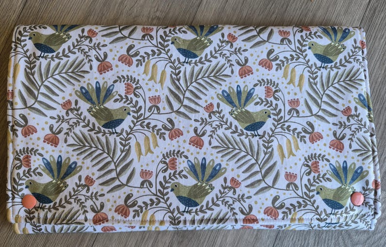 May include: A rectangular, folded fabric item featuring a repeating pattern of birds, flowers, and foliage in green, blue, and orange on a white background. Secured with two orange buttons.