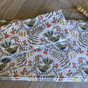 May include: White fabric featuring a repeating pattern of birds, flowers, and foliage in green, blue, and orange. The fabric is folded, possibly a blanket or bedding set. A sprig of wheat is in the background. The measurements are in centimeters.