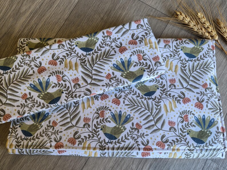 May include: White fabric featuring a repeating pattern of birds, flowers, and foliage in green, blue, and orange. The fabric is folded, possibly a blanket or bedding set. A sprig of wheat is in the background. The measurements are in centimeters.