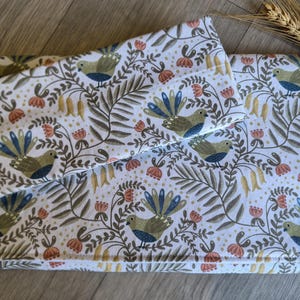 May include: A set of white fabric items featuring a repeating pattern of birds, flowers, and foliage in green, blue, and orange. The items appear to be a pillowcase and a folded blanket or cover.