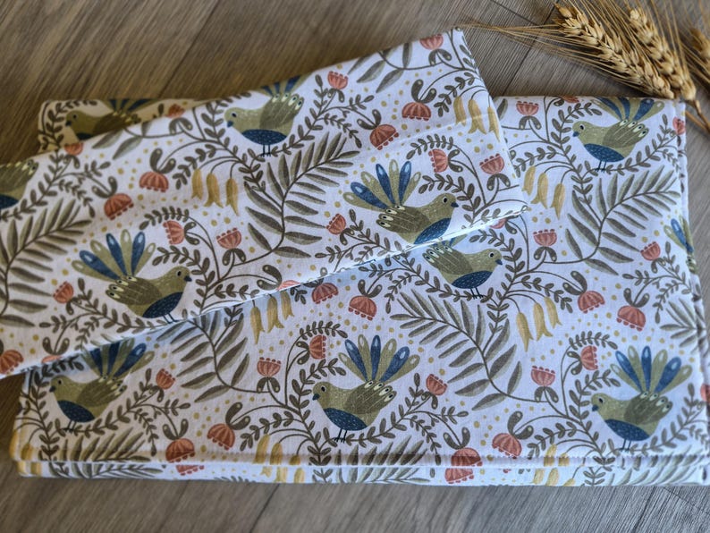 May include: A set of white fabric items featuring a repeating pattern of birds, flowers, and foliage in green, blue, and orange. The items appear to be a pillowcase and a folded blanket or cover.