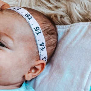 May include: A baby's head is measured with a white measuring tape. The tape, marked with black numbers and lines, is wrapped around the baby's head. The baby is lying on a white blanket, with a fluffy, light-coloured surface in the background.