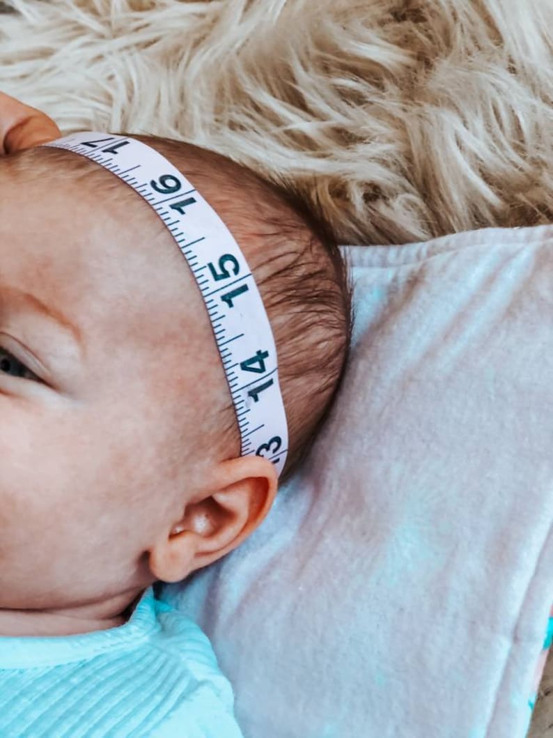 May include: A baby's head is measured with a white measuring tape. The tape, marked with black numbers and lines, is wrapped around the baby's head. The baby is lying on a white blanket, with a fluffy, light-coloured surface in the background.
