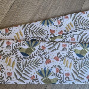 May include: White fabric featuring a repeating pattern of birds, flowers, and foliage in green, blue, orange, and yellow. The fabric is folded, revealing the detailed design. The overall theme is nature and botanical.