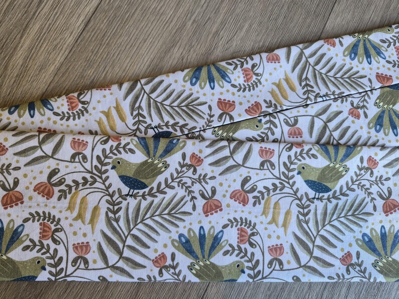 May include: White fabric featuring a repeating pattern of birds, flowers, and foliage in green, blue, orange, and yellow. The fabric is folded, revealing the detailed design. The overall theme is nature and botanical.