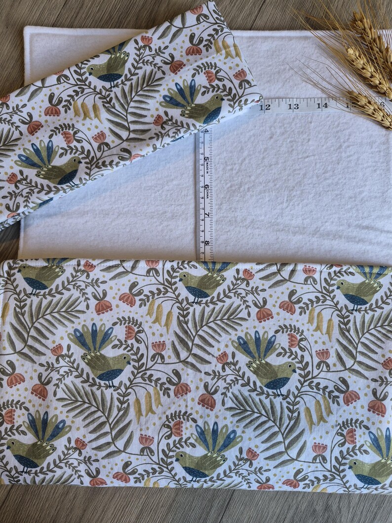 May include: White changing mat cover with a colorful bird and floral pattern. The pattern includes birds with blue and green feathers, red flowers, and green leaves. A ruler shows the mat is approximately 35 cm long.