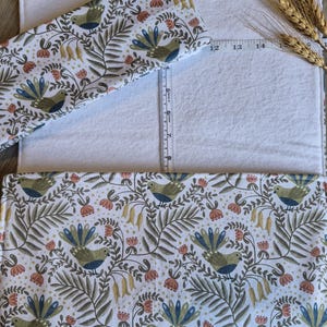 May include: A white rectangular fabric with a patterned fabric overlay featuring birds, flowers, and foliage in shades of green, blue, and peach. A measuring tape is placed across the fabric, and a smaller fabric piece is on top. The fabric is approximately 35.5 cm long.
