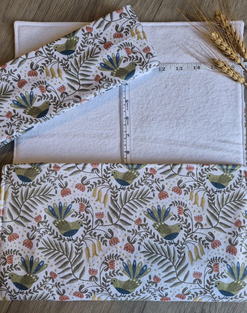May include: A white rectangular fabric with a patterned fabric overlay featuring birds, flowers, and foliage in shades of green, blue, and peach. A measuring tape is placed across the fabric, and a smaller fabric piece is on top. The fabric is approximately 35.5 cm long.