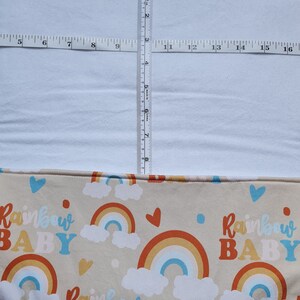 May include: A beige fabric with a repeating pattern of rainbows, clouds, and the text "Rainbow Baby". The fabric is folded in half and has a white fabric layer on top.