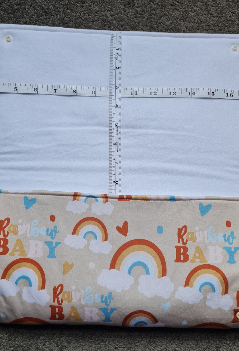 May include: A beige fabric with a repeating pattern of rainbows, clouds, and the text "Rainbow Baby". The fabric is folded in half and has a white fabric layer on top.