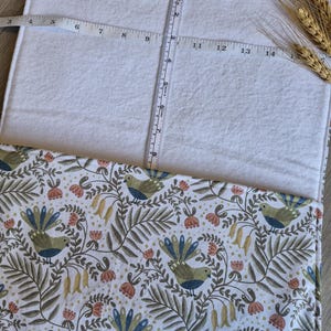 May include: A white rectangular fabric with a patterned border featuring birds, flowers, and foliage in shades of green, blue, and peach. A measuring tape shows the fabric is approximately 25 cm wide. Wheat stalks are in the upper right corner.