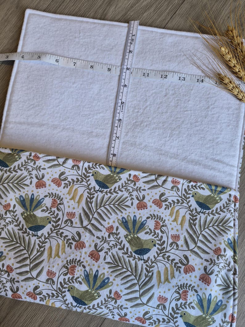 May include: A white rectangular fabric with a patterned border featuring birds, flowers, and foliage in shades of green, blue, and peach. A measuring tape shows the fabric is approximately 25 cm wide. Wheat stalks are in the upper right corner.