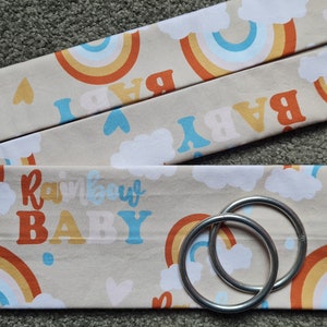 May include: Two beige fabric straps with a rainbow and cloud pattern and the text "Rainbow Baby". The straps are attached to two silver rings.