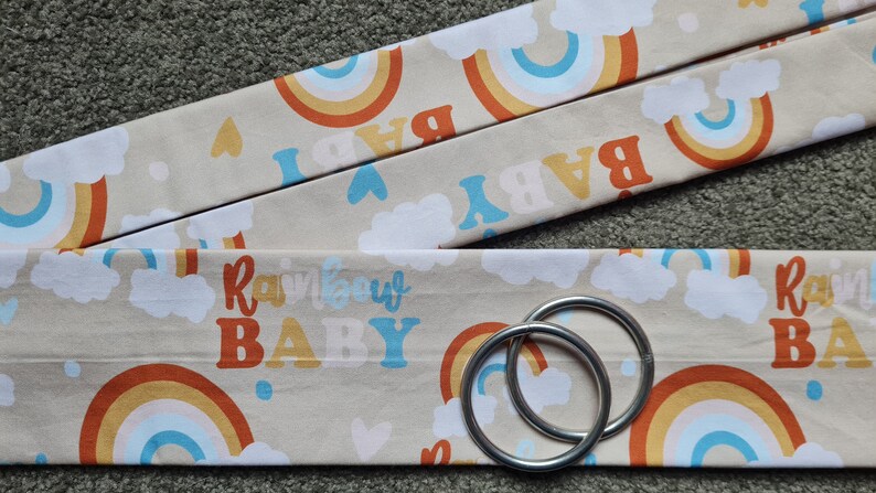 May include: Two beige fabric straps with a rainbow and cloud pattern and the text "Rainbow Baby". The straps are attached to two silver rings.