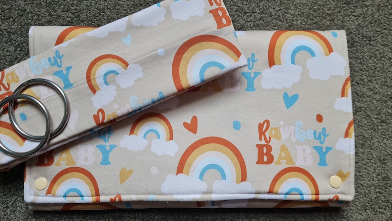 May include: A set of two beige fabric burp cloths with a rainbow and cloud pattern. The larger cloth has the text "Rainbow Baby" printed on it. The smaller cloth has two metal rings attached to it.