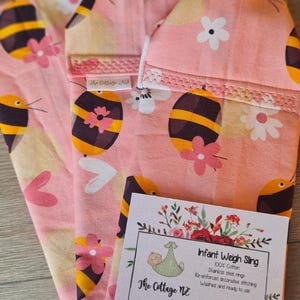 May include: Pink infant weigh sling with a bee and flower pattern. The sling is made of 100% cotton and features stainless steel rings. The tag reads "Infant Weigh Sling" and includes the shop's name and website.