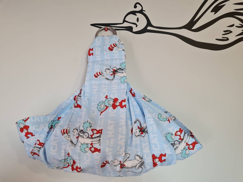 May include: A blue baby sling with a white background and a pattern of the Cat in the Hat characters. The sling is hanging from a hook.