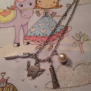 May include: A silver necklace with a charm of a tooth with wings, a charm of a tube of toothpaste, and a pearl charm. The necklace is on a blue and white fabric with a cartoon unicorn and princess.
