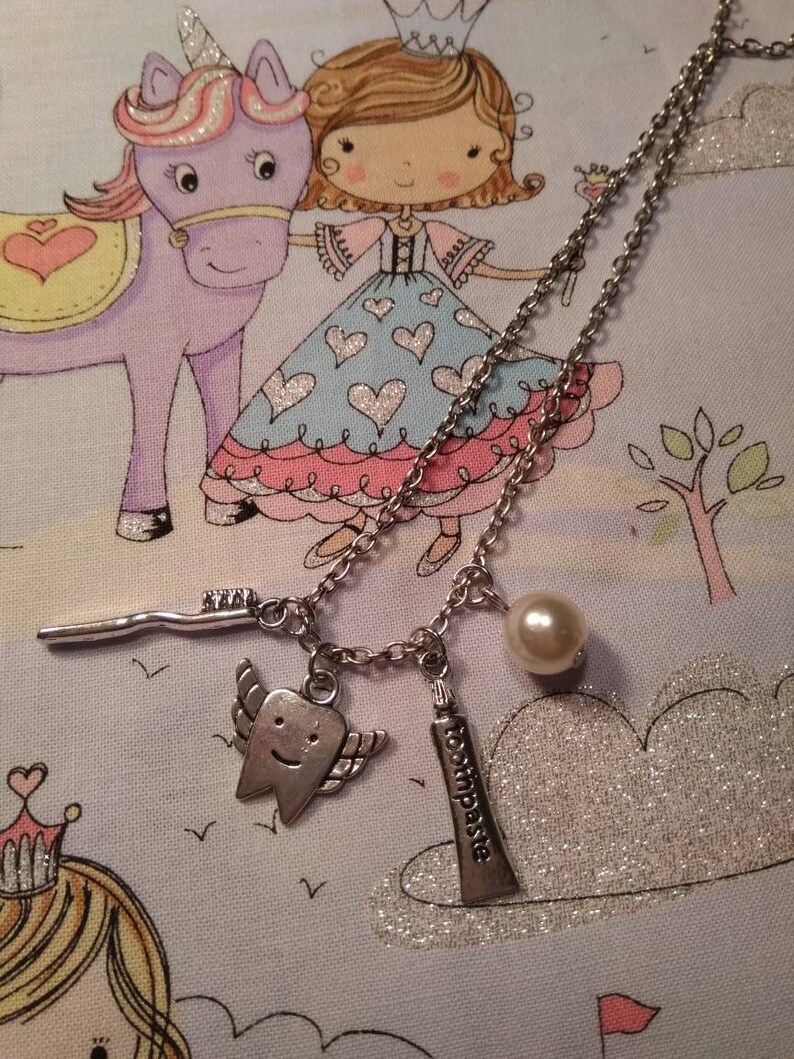 May include: A silver necklace with a charm of a tooth with wings, a charm of a tube of toothpaste, and a pearl charm. The necklace is on a blue and white fabric with a cartoon unicorn and princess.
