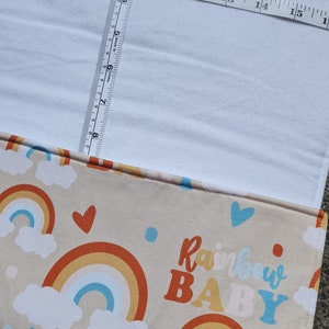 May include: A beige fabric with a rainbow and cloud pattern. The fabric has the text "Rainbow Baby" in orange, yellow, and blue letters. The fabric is measured with a tape measure.