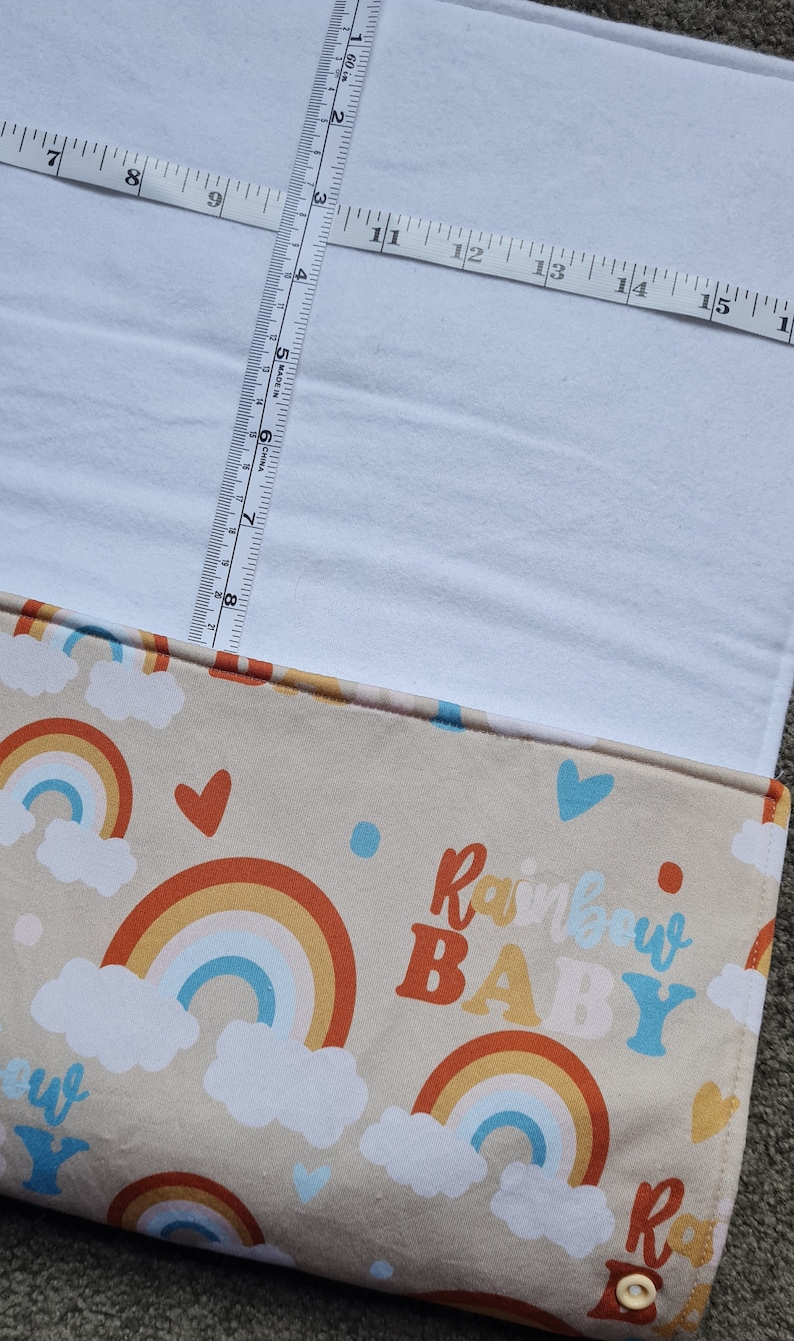 May include: A beige fabric with a rainbow and cloud pattern. The fabric has the text "Rainbow Baby" in orange, yellow, and blue letters. The fabric is measured with a tape measure.
