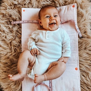 May include: A baby in a white ribbed onesie smiles on a pink mat with a measuring tape. The baby is lying on a fluffy, light brown rug. The onesie has long sleeves and three buttons at the neckline.
