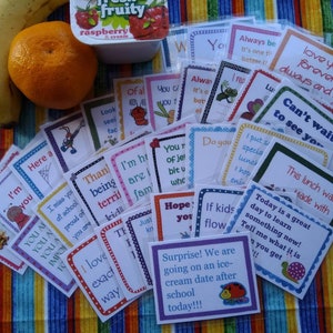 Può includere: Un assortimento colorato di piccole carte con messaggi incoraggianti per i bambini, come "Sono orgoglioso di te" e "Sei una stella". Le carte sono disposte su una tovaglia a righe multicolori con una banana e un'arancia sullo sfondo.