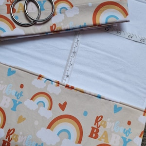 May include: A beige fabric baby changing mat with a rainbow and cloud pattern. The text "Rainbow Baby" is repeated throughout the fabric. The mat has two metal rings attached to the top flap.
