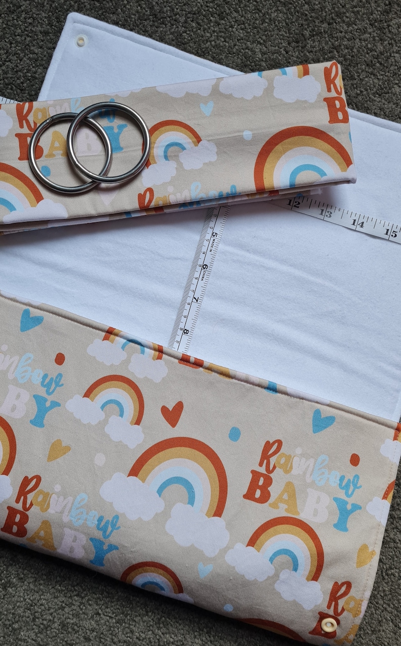 May include: A beige fabric baby changing mat with a rainbow and cloud pattern. The text "Rainbow Baby" is repeated throughout the fabric. The mat has two metal rings attached to the top flap.