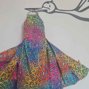May include: A baby carrier with a vibrant, colourful pattern. The fabric displays a rainbow of hues, including orange, yellow, green, blue, and purple, with a black and white swirling design. A silver metal ring is at the top.