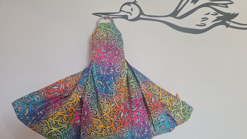 May include: A baby carrier with a vibrant, colourful pattern. The fabric displays a rainbow of hues, including orange, yellow, green, blue, and purple, with a black and white swirling design. A silver metal ring is at the top.