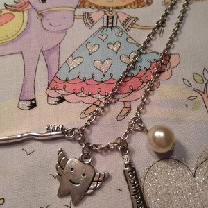 May include: A silver chain necklace with three charms: a tooth with wings, a tube of toothpaste that says "toothpaste", and a pearl. The necklace is on a light blue and pink fabric background with a cartoon drawing of a princess and a unicorn.