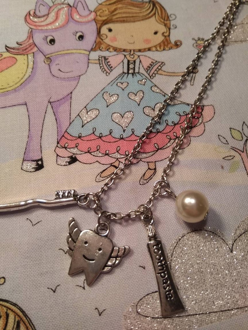 May include: A silver chain necklace with three charms: a tooth with wings, a tube of toothpaste that says "toothpaste", and a pearl. The necklace is on a light blue and pink fabric background with a cartoon drawing of a princess and a unicorn.