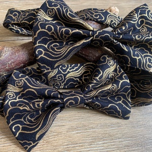 May include: Two black bow ties with a gold cloud pattern. The bow ties are made of fabric and are tied together.