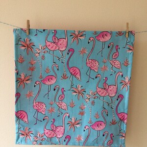 May include: A blue tea towel with a pink flamingo pattern. The flamingos are in various poses, some with their necks curved and others with their wings spread. The towel is decorated with palm trees and a white background.